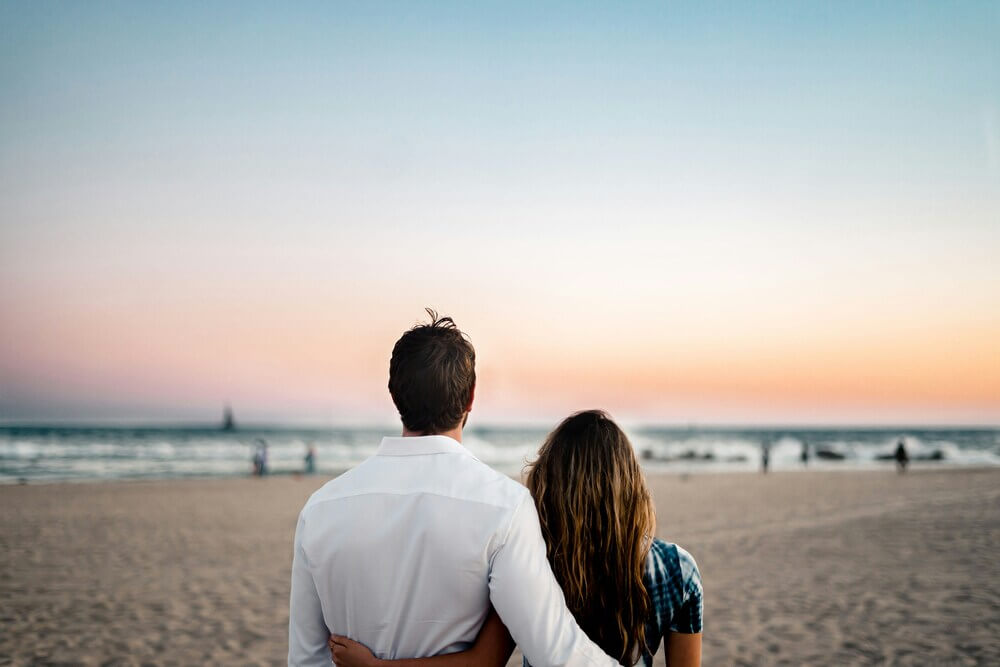 Couple embracing and watching the sunset over the ocean on a sandy beach.